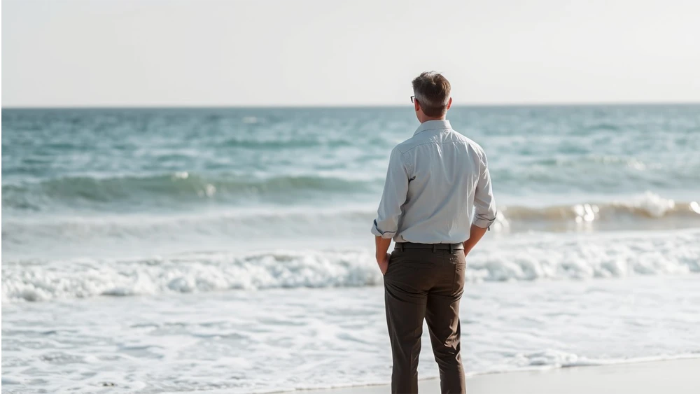 Un hombre vestido con pantalones y camisa en la playa observando el mar con las manos en los bolsillos